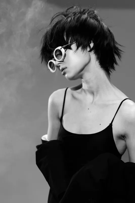 Black and white portrait of a young woman with short hair wearing round glasses and a black top, showing a thoughtful and stylish mood.