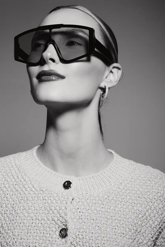 Stylish white woman with pale skin wearing oversized sunglasses and a knitted sweater poses confidently against a plain background in black and white.