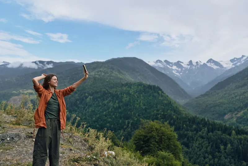 Mountain landscape with a person taking a selfie on a ridge, surrounded by forested slopes and blue sky, conveying outdoor adventure, exploration, and natural freedom.