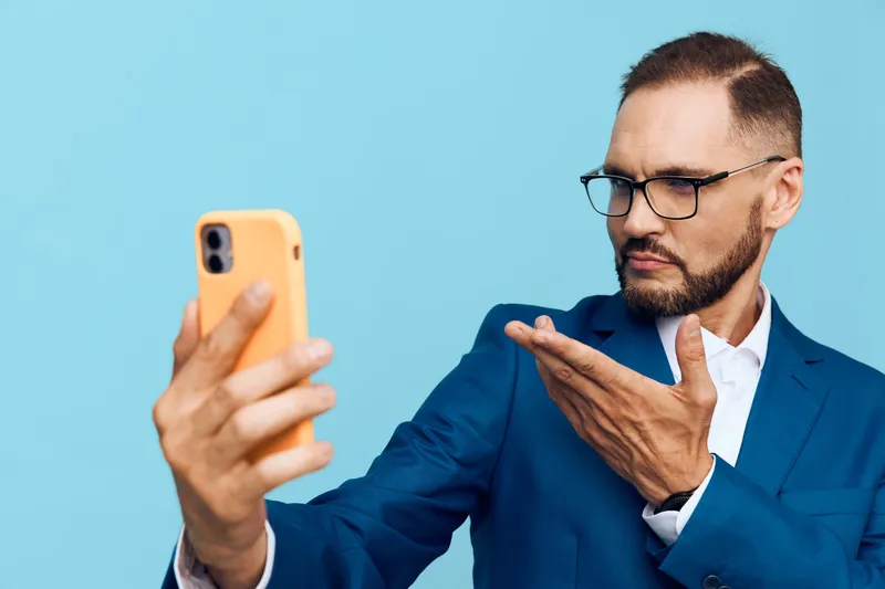 A business professional man on a vibrant blue background taking a selfie with a smartphone, displaying thoughtful, focused emotions and confident posture.