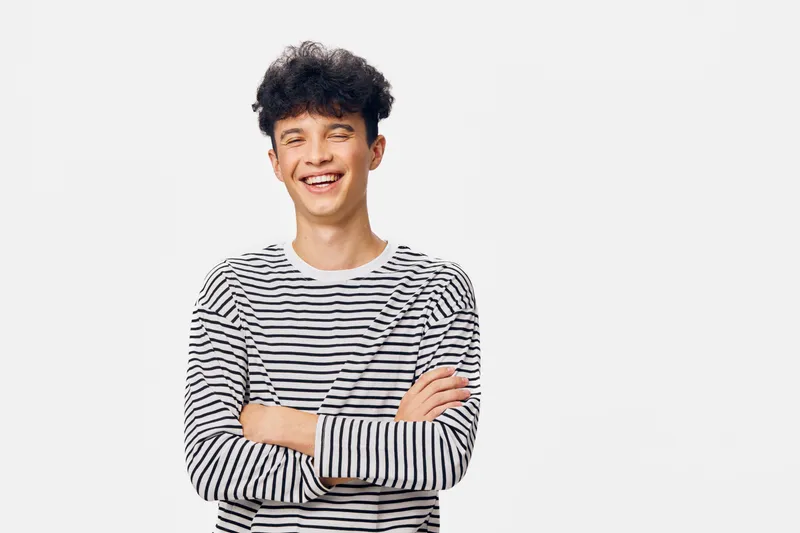 happy young man with curly hair wearing a striped shirt, smiling with crossed arms against a plain white background, casual and cheerful mood