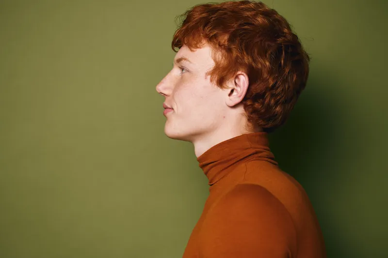 Profile portrait of young man with red hair wearing orange turtleneck standing against green background in studio, side view with calm expression and natural lighting.