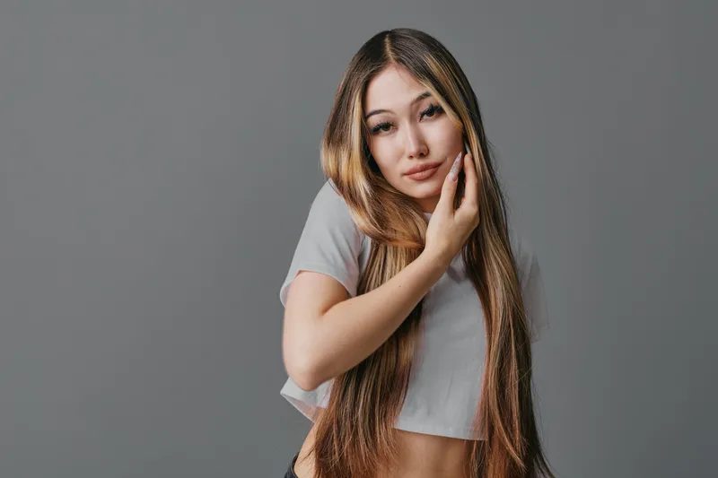 Young woman with long straight hair and casual outfit posing with hand on face against neutral background for beauty and fashion concept.