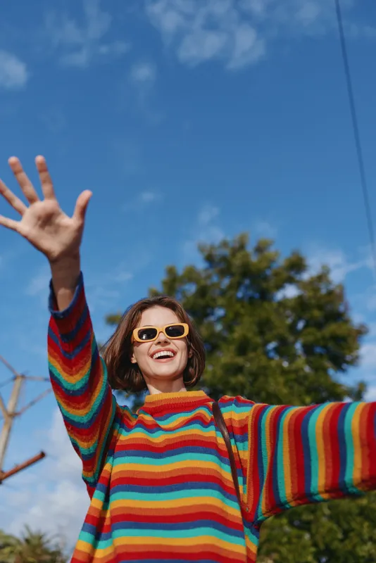Woman in a rainbow striped sweater smiling and waving outdoors under blue sky, wearing yellow sunglasses and casual outfit with backpack, joyful travel lifestyle portrait in suburb.
