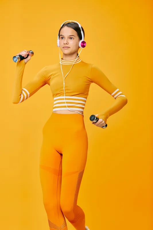 Young woman fitness athlete in matching orange workout set holds small dumbbells and wears headphones, exercising with confident posture against a vibrant solid orange background.