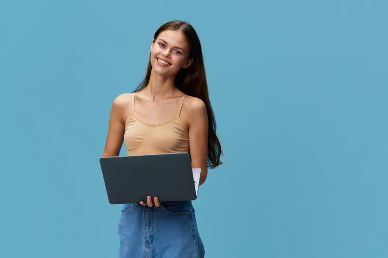 smiling woman holding laptop, casual clothing, blue background, young adult woman working, technology usage, happy expression