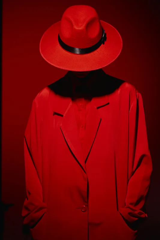 Stylish figure dressed in red coat and matching hat poses with face hidden by hat, creating a mysterious fashion and beauty concept with bold, monochrome color.