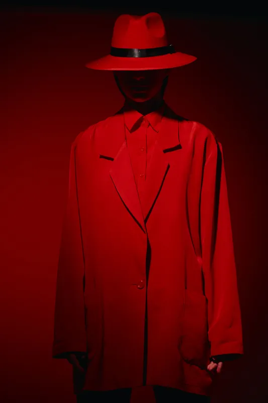 Woman in oversized red blazer and wide red hat with black band standing in dramatic lighting on red background. Fashion portrait with creative bold color concept.