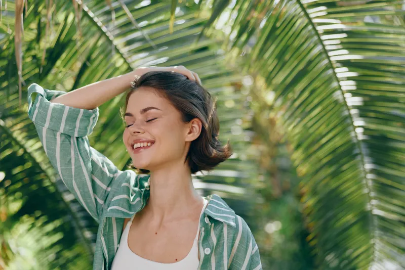 Woman stands outdoors in a sunlit tropical setting, smiling with her hand in her hair, wearing a striped shirt, surrounded by palm leaves, radiating relaxed confidence and natural joy.