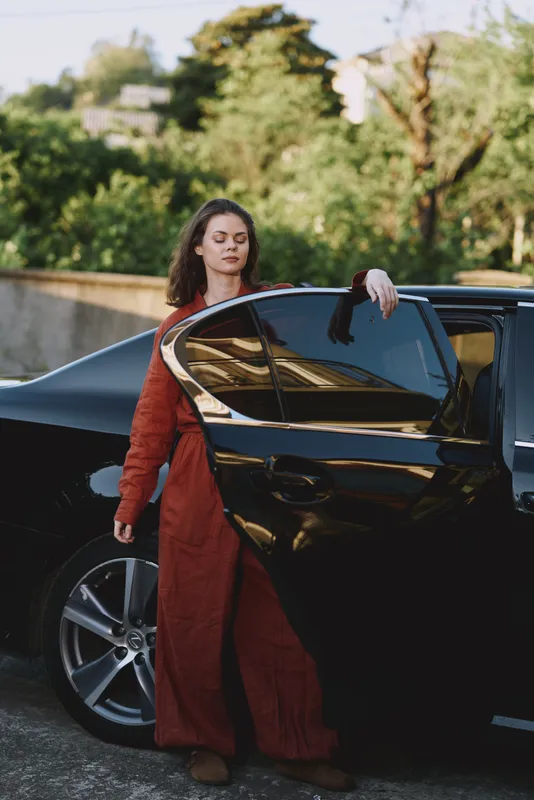 A stylish woman in a red coat exits a sleek black car on a sunlit street, embodying confident, everyday fashion and modern lifestyle