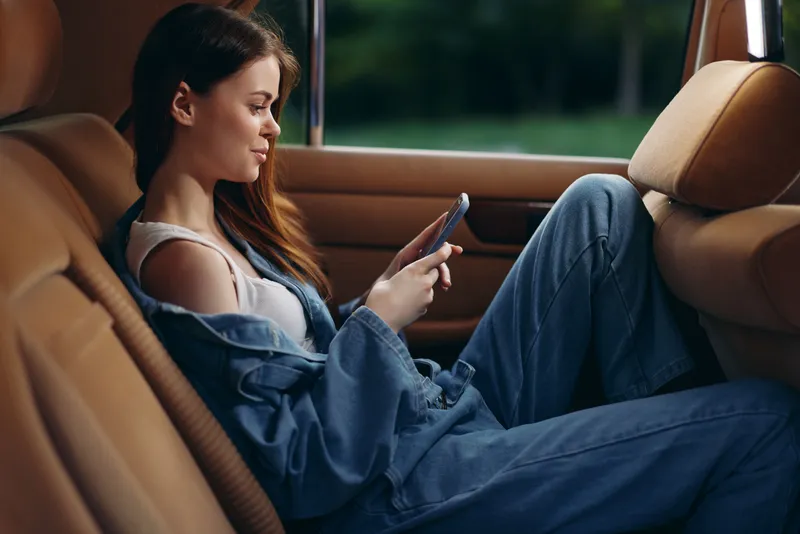 Young woman in casual denim outfit using smartphone in a car, expressing relaxed emotions with a vibrant green background, perfect for lifestyle themes.