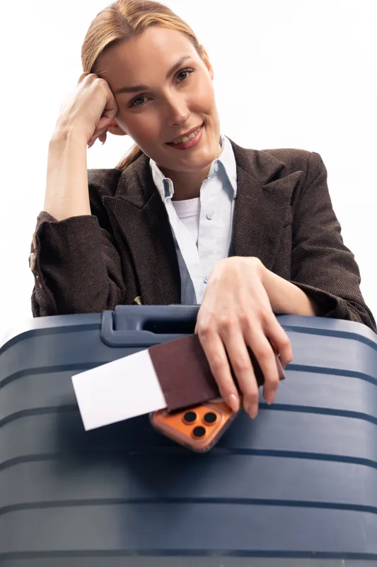 Smiling woman in business outfit leans on blue suitcase holding passport and phone. White background. Concept of travel, business trip, vacation, and modern lifestyle.