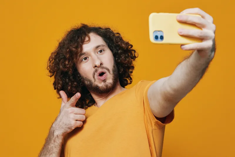 Cheerful young man with curly hair taking a selfie against a vibrant yellow background, showcasing joy and self expression in a casual orange t shirt