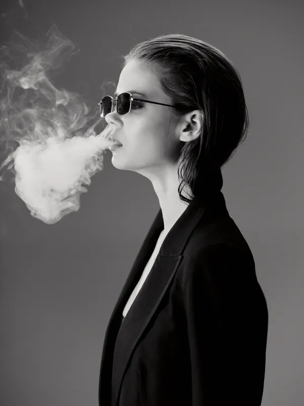 Black and white portrait of a woman smoking with sunglasses and a stylish jacket, exhaling a thick cloud of smoke against a gray background.