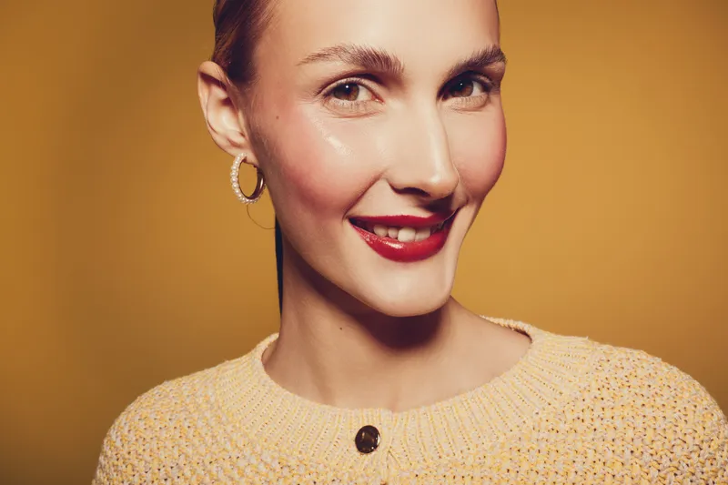 Smiling woman with red lipstick and hoop earrings wears yellow knitted sweater on yellow background. Close-up portrait with natural makeup and confident expression for beauty concept.