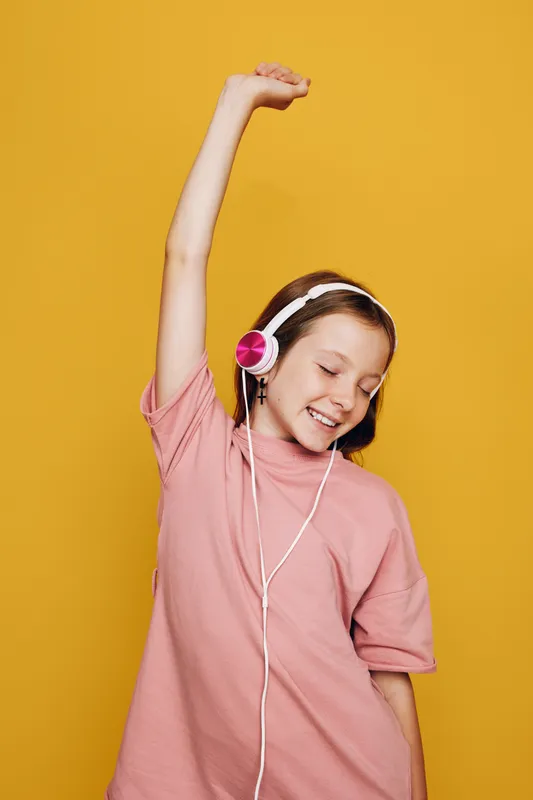 Happy girl in pink t-shirt wearing headphones enjoys music with eyes closed and arm raised. Yellow background. Concept of joy, relaxation, and youth lifestyle.