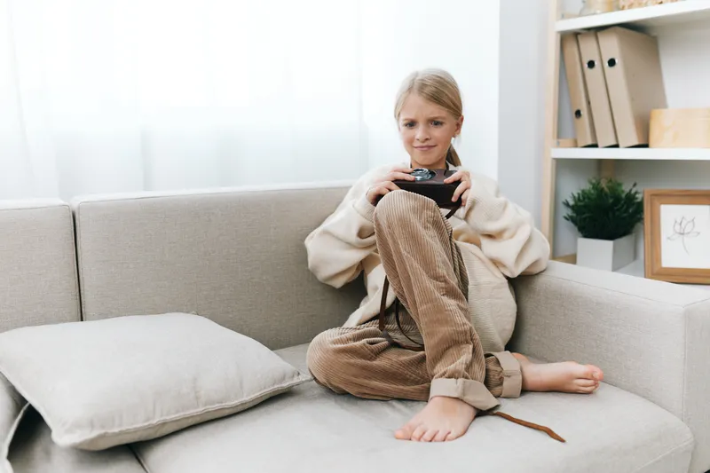 Young girl sits on a cozy sofa, holding a camera, natural light bathes the room. Casual outfit, soft textures, and a calm, focused mood capture her playful curiosity.