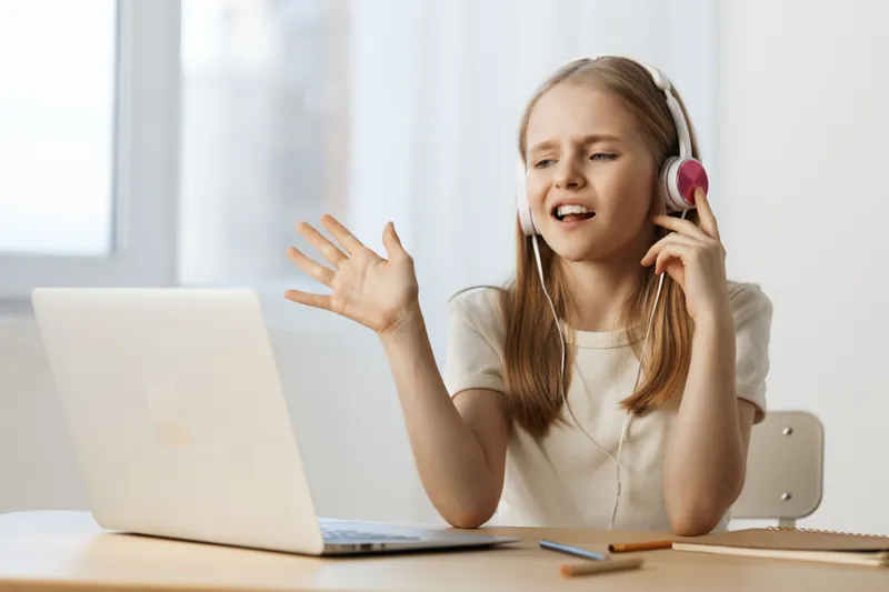 Young girl with headphones engaging in an online class, showcasing excitement and concentration while sitting at her desk with a laptop, in a bright modern room