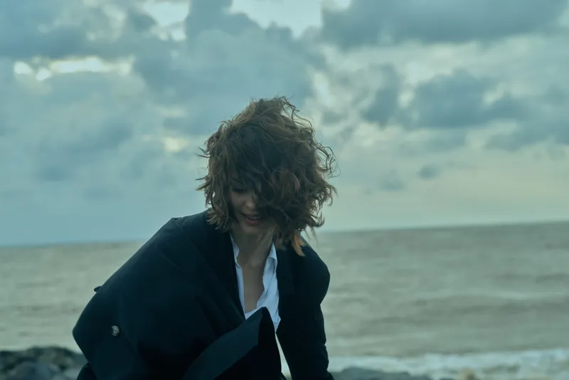 Windy coastal scene with an individual in a dark jacket by the ocean, as waves crash and clouds roll over the horizon, creating a moody seascape