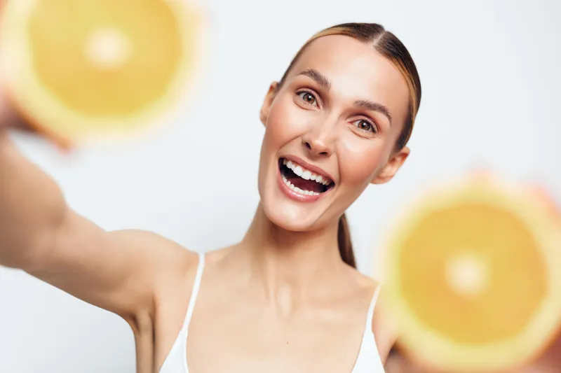 Happy young woman with ponytail holds two halves of fresh orange fruit near camera smiles brightly isolated on white background healthy lifestyle concept.