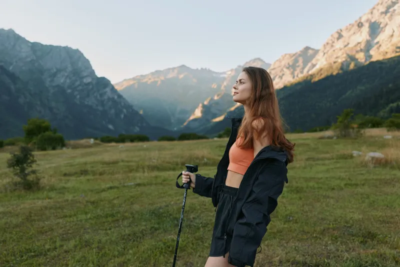 A young woman stands in a mountain valley, holding a trekking pole. She wears a lightweight jacket and sportswear, looking into the distance with a calm, focused expression.