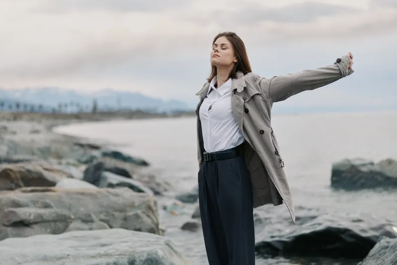 Young woman in stylish coat stands on rocky beach with arms outstretched, eyes closed, enjoying fresh sea breeze and calm atmosphere. Concept of freedom, relaxation, and nature connection.