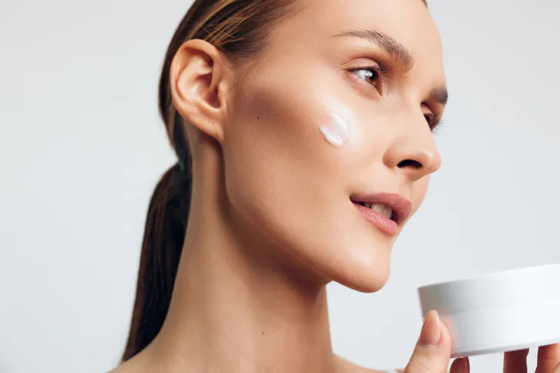 A young brunette woman applies cream to her face in a studio closeup, capturing a skincare moment with soft lighting, smooth skin, and a calm, focused expression