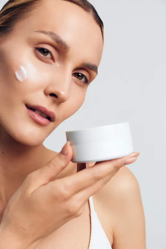 Young woman with clear skin holding white cream jar applying moisturizer on cheek. Close-up portrait on light background. Concept of skincare, beauty, and daily self-care routine.