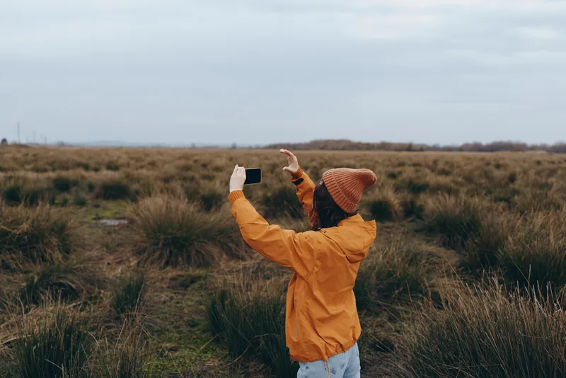 Woman lifestyle, outdoor field, nature walk through autumn grass, orange jacket and knit hat, arms raised, cloudy sky overhead, recreation and freedom, soft daylight, calm mood, today