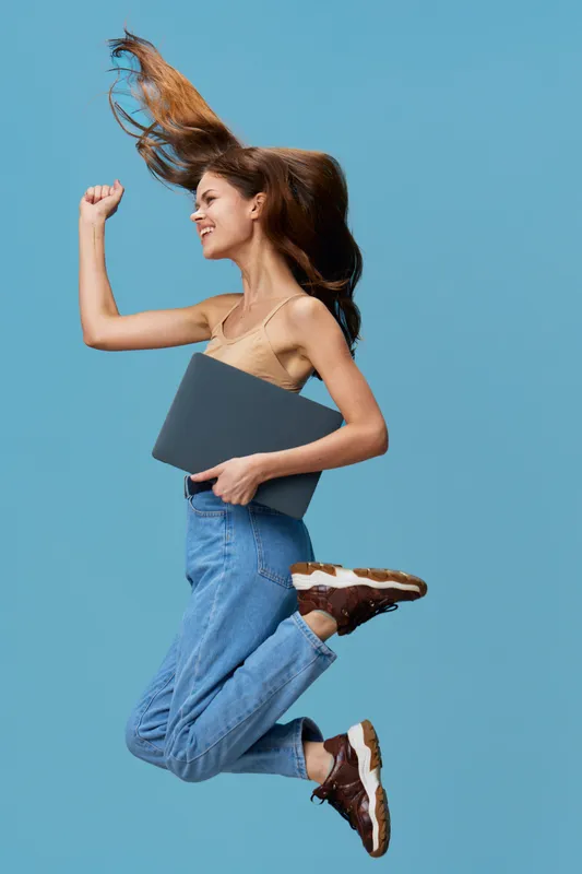 Happy young woman jumping energetically holding a laptop, wearing casual beige top and jeans against blue background, showing excitement and freedom.