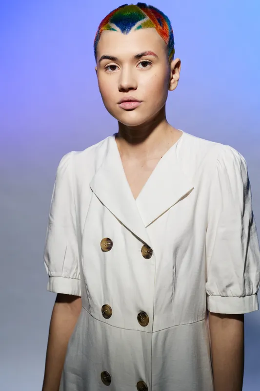 young woman with colorful short hair wearing white vintage dress standing against gradient purple background, minimal expression, contemporary fashion style.