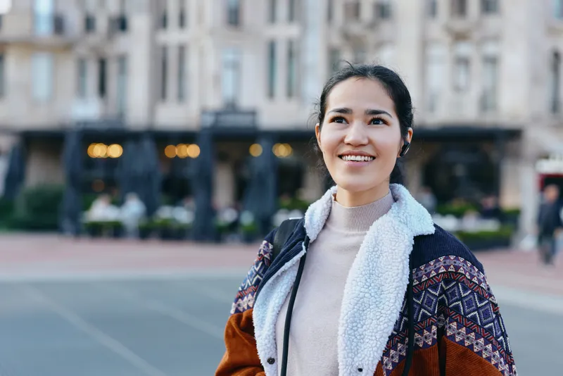 Smiling woman in outdoor street scene wearing jacket, cardigan and scarf. Casual urban fashion, daylight ambiance, candid moment capturing warmth, confidence and style amid storefronts and