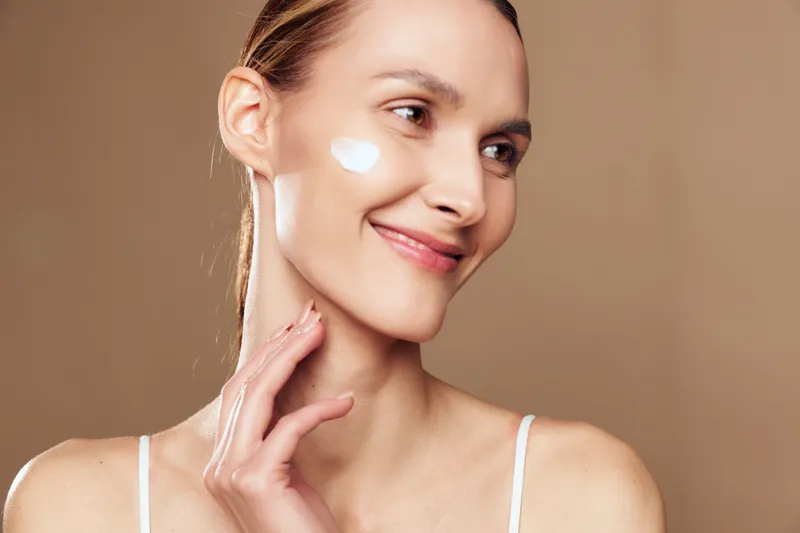 Woman with smile applying face cream, close up portrait, soft studio lighting on beige background, skincare and beauty routine, wellness self care concept, healthy glowing skin