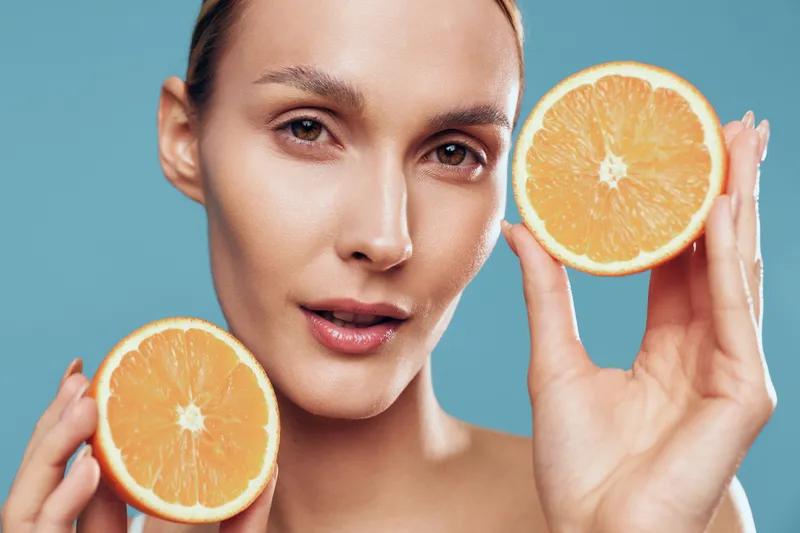 Close-up of a woman holding orange halves, vibrant citrus colors, smooth skin, refreshing skincare moment, bright blue backdrop, natural lighting, confident expression, studio setting, summery vibe
