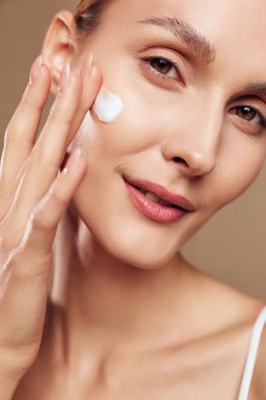 Close-up of smiling woman applying white face cream on cheek, natural skin care routine on beige background, healthy glowing skin, beauty and wellness concept with soft lighting.