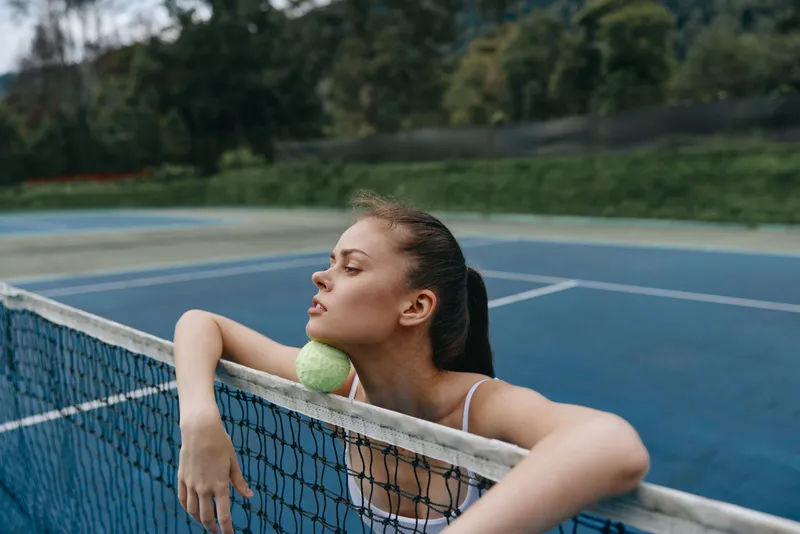 Young female tennis player resting on the net with a ball, showcasing determination and focus on a sunny day at a sports court