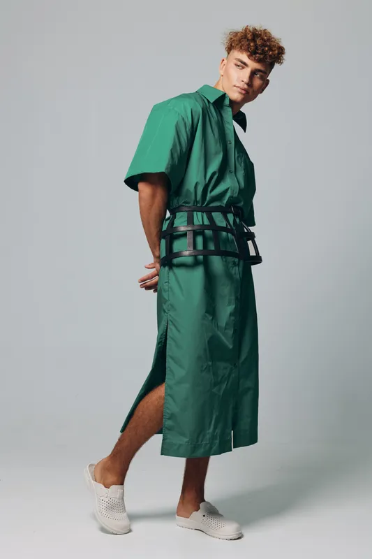 Young man with curly hair wearing green oversized dress and white slip-on shoes posing in studio with hands behind back and serious expression on gray background.