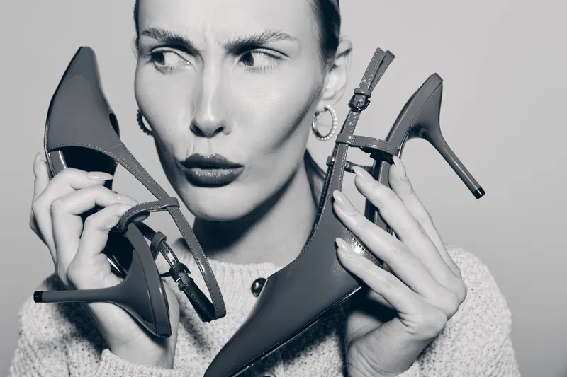 White woman with light skin examines two high heel shoes with confused expression. Close-up in black and white. Concept of fashion choice and style decision.