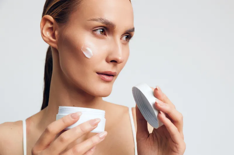 Young woman applies cream on her cheek from a jar, close up skincare moment, soft lighting, clean background, healthy glow, calm expression, personal care