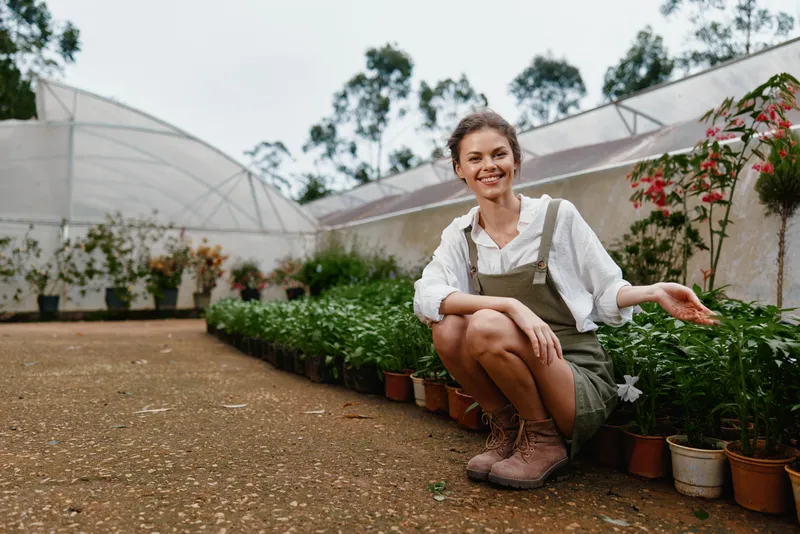 Young woman, smiling and kneeling in a greenhouse surrounded by potted plants, wearing casual clothing, embodying the concept of sustainable gardening
