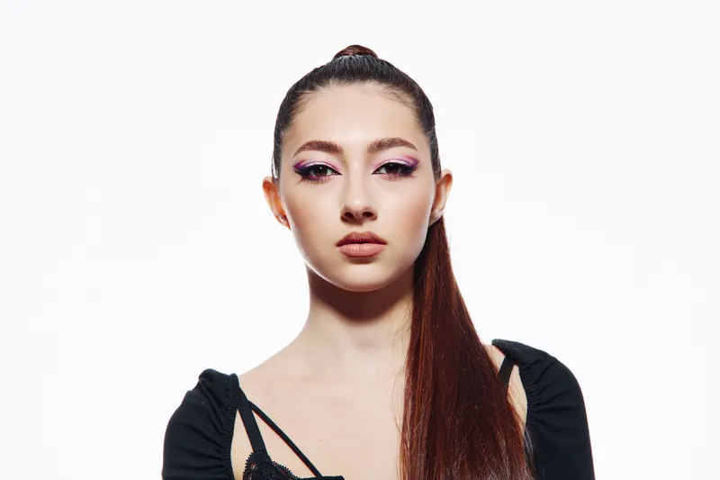 Young woman with long brown hair in sleek ponytail wearing black clothes poses with creative purple eye makeup against a white studio background. Concept of beauty and fashion.