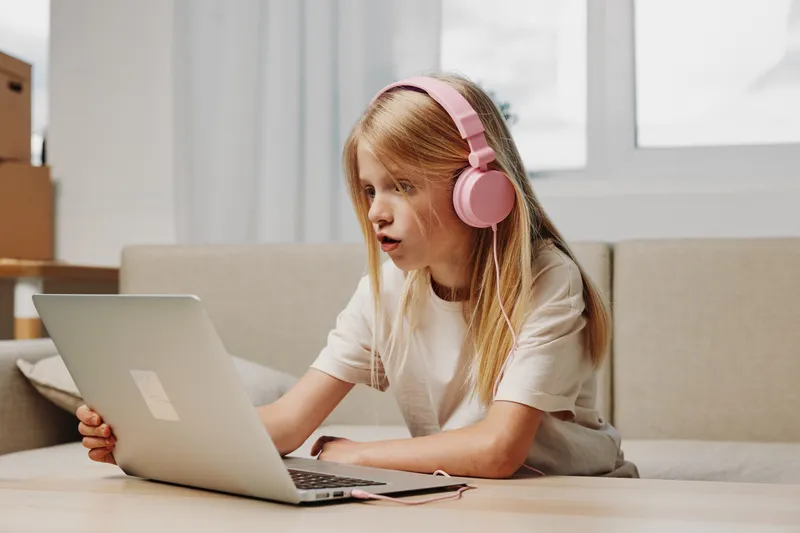 Curious girl with pink headphones uses laptop at home, focused on screen. Natural light brightens modern cozy living room interior.
