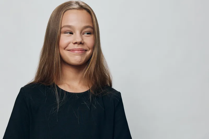 Girl with long hair smiling and looking to side in black shirt on plain light background, youth, happiness, casual portrait with natural expression and positive mood.