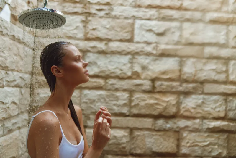 woman showering outdoors under rain shower with wet hair against stone wall, serene expression, natural light, summer wellness scene