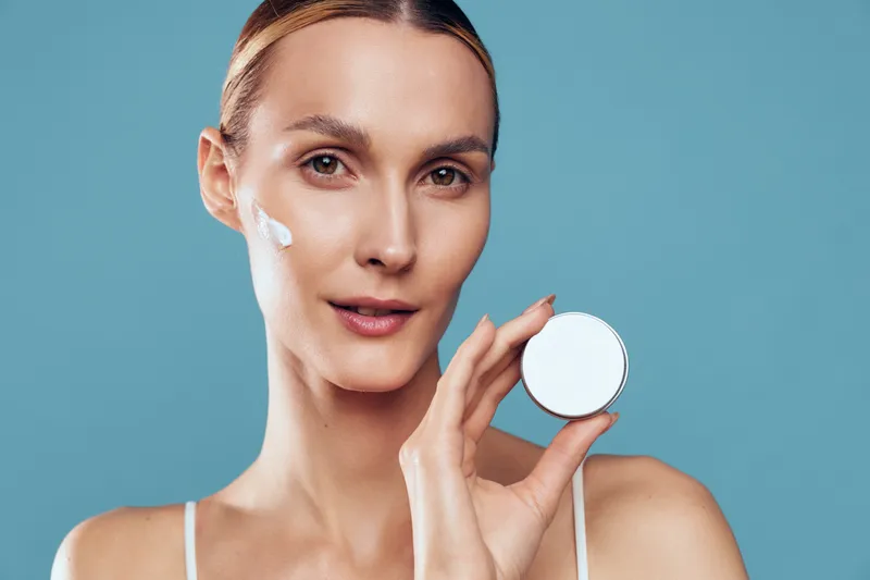 Smiling woman with a white cream applied on her cheek, close-up portrait against a blue background, skincare, beauty, and selfcare concept, soft lighting creates a gentle glow