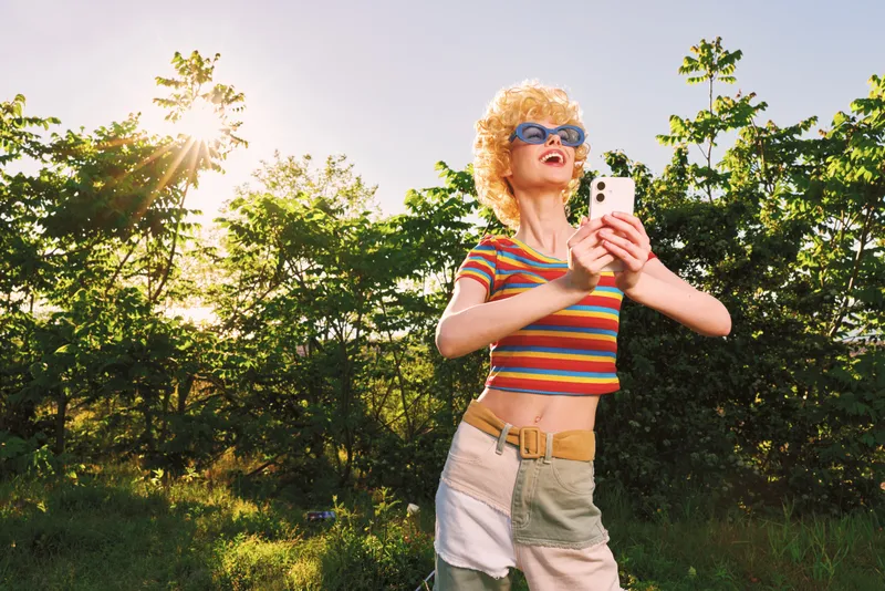 Cheerful woman outdoors in retro style, striped crop top and sunglasses, golden sunlight, relaxed pose, capturing moment on phone, lush trees in background, warm natural tones against a clear sky