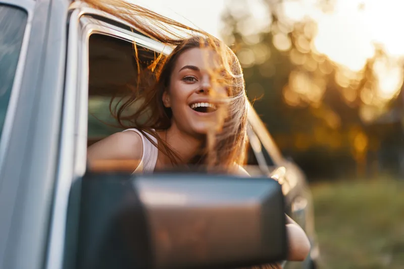 Happy young woman enjoying a road trip with the wind in her hair, wearing casual clothing, in a warm sunny setting, perfect for adventure and travel concepts.