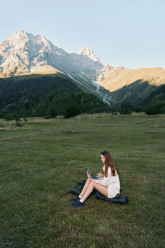 A young woman sits alone in a grassy meadow, using a laptop in a mountain landscape. She appears focused and relaxed, blending work with nature in a serene outdoor setting.
