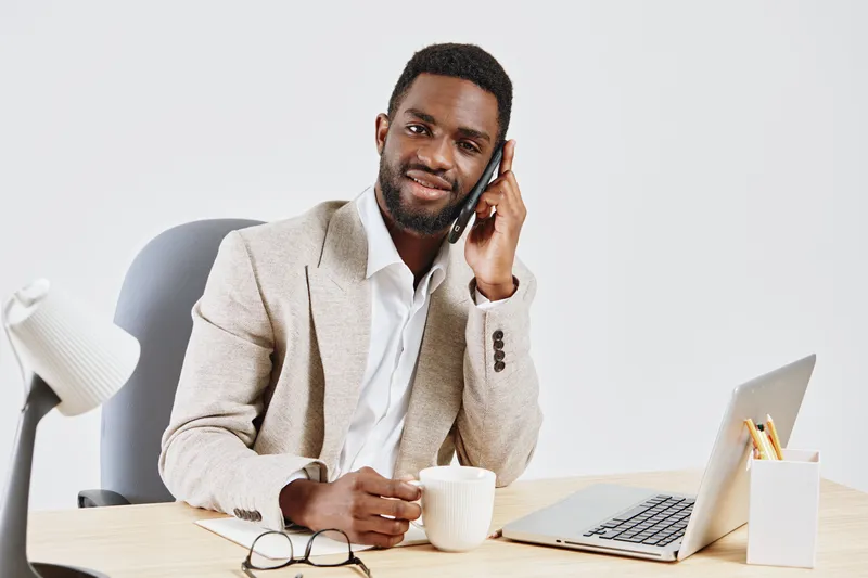 Professional businessman with dark skin talking on mobile phone, smiling, seated at modern office desk with laptop, coffee mug, glasses, isolated on plain white background. Business communication conc