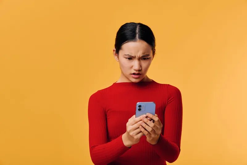 Woman with angry face holding smartphone and looking at screen for negative message or bad news on yellow background. Emotional person, technology and communication concept in studio.